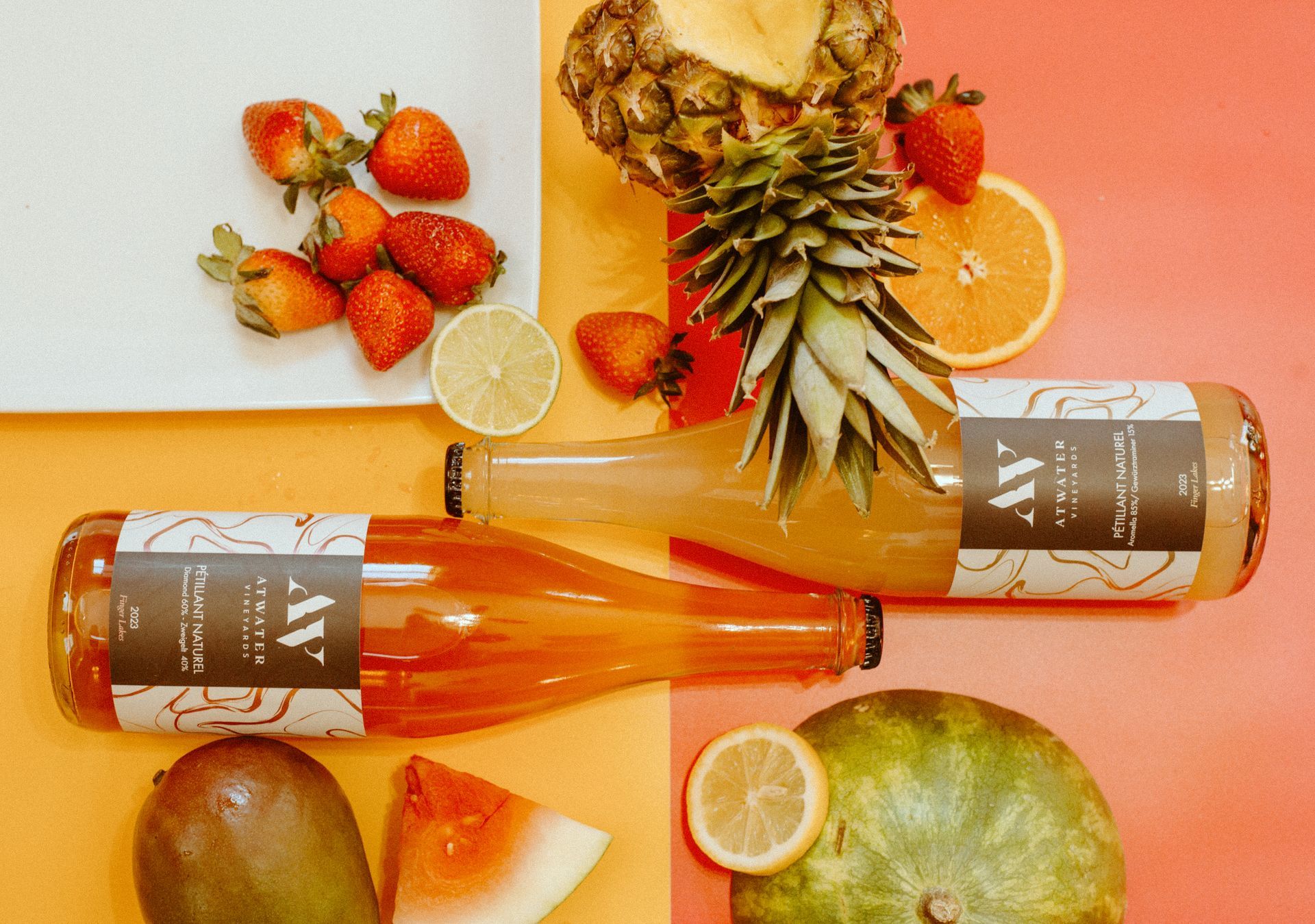 Two bottles of fruit-flavored drinks surrounded by pineapples, strawberries, mango, oranges, and lemons on a two-tone mat.
