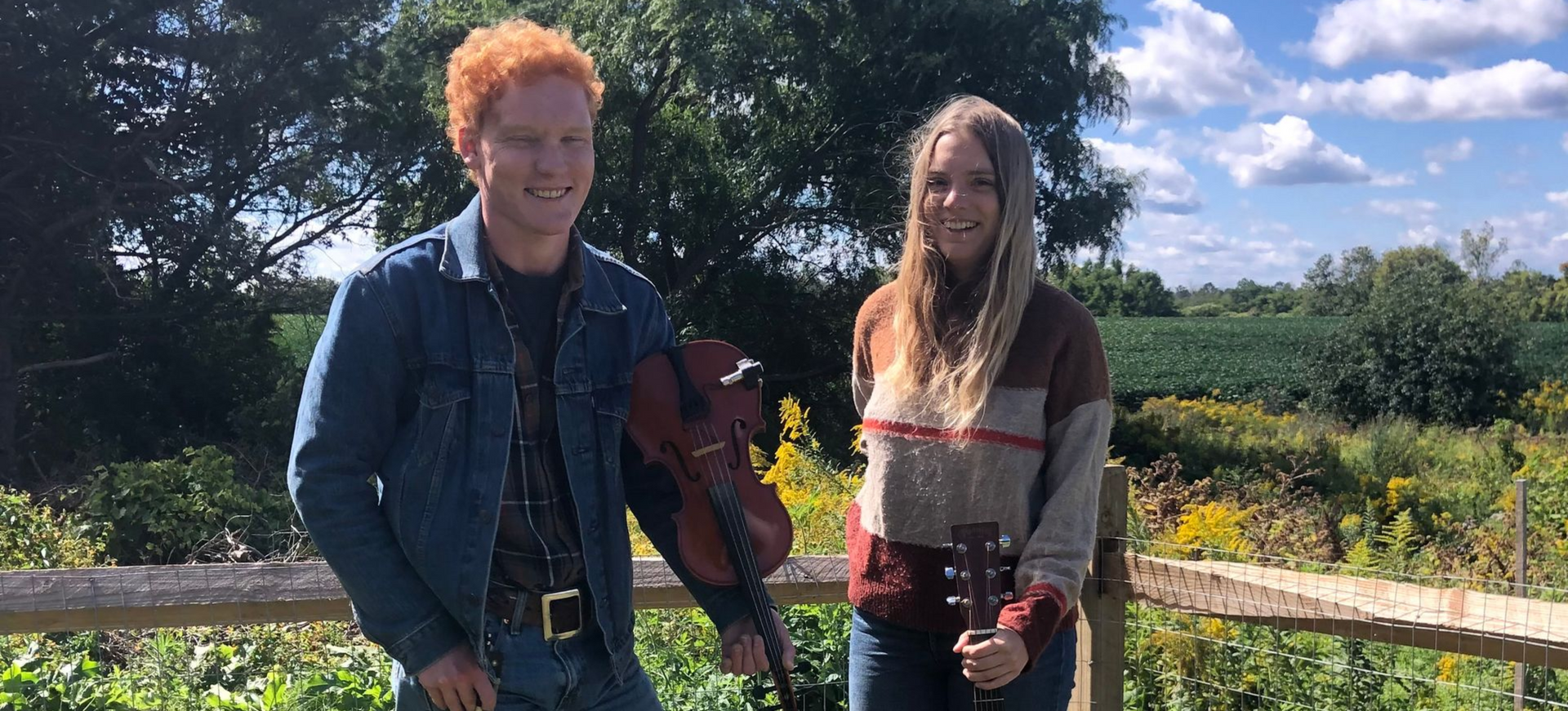 Two musicians smile outdoors: a redhead with a violin, a woman with a guitar, in front of a field on a sunny day.