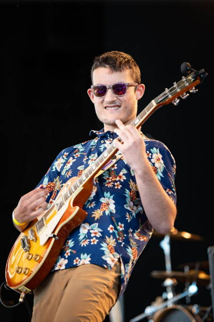 A person in sunglasses and a floral shirt plays an electric guitar while standing on a stage against a dark background.
