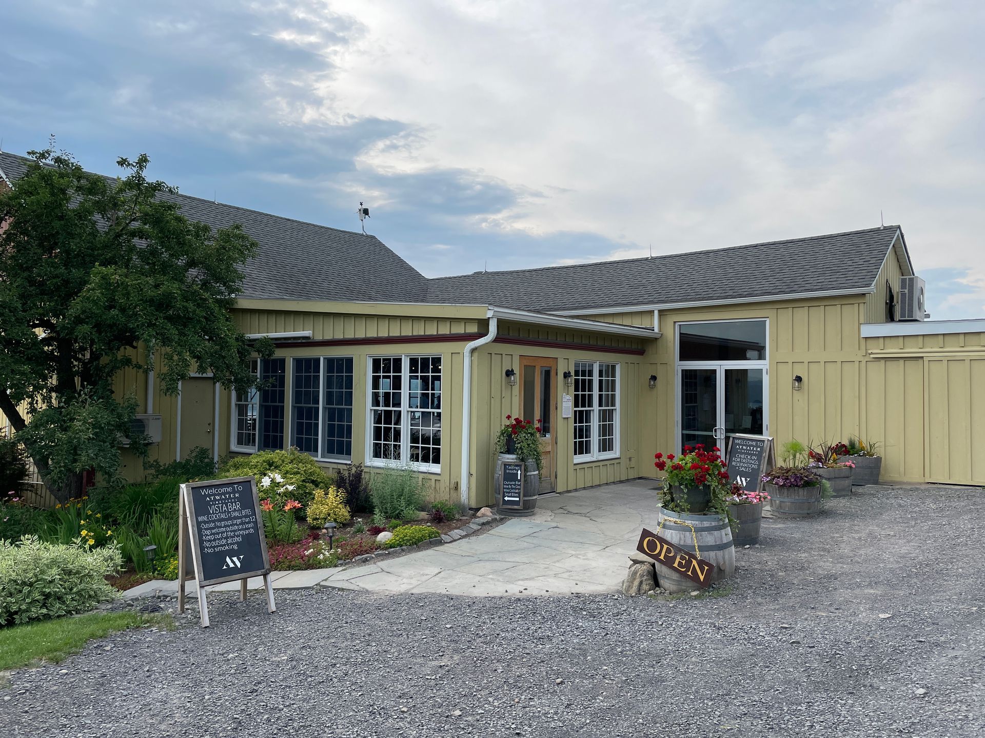 A light yellow winery with a gravel parking area, a chalkboard sign, and potted flowers under a cloudy sky.