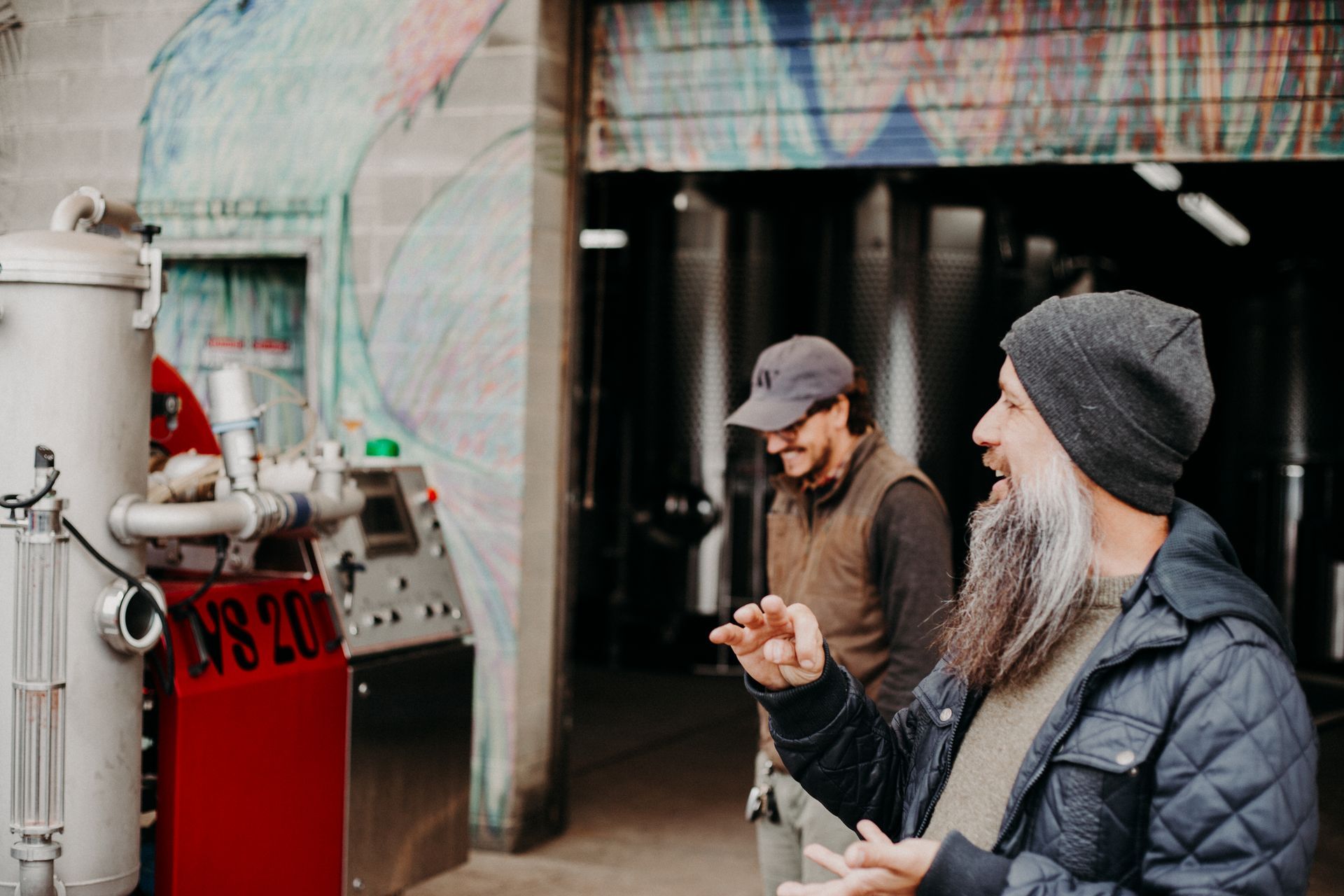 Two men in a wine making facility; one with a long beard gestures, the other smiles. Gray and red equipment.