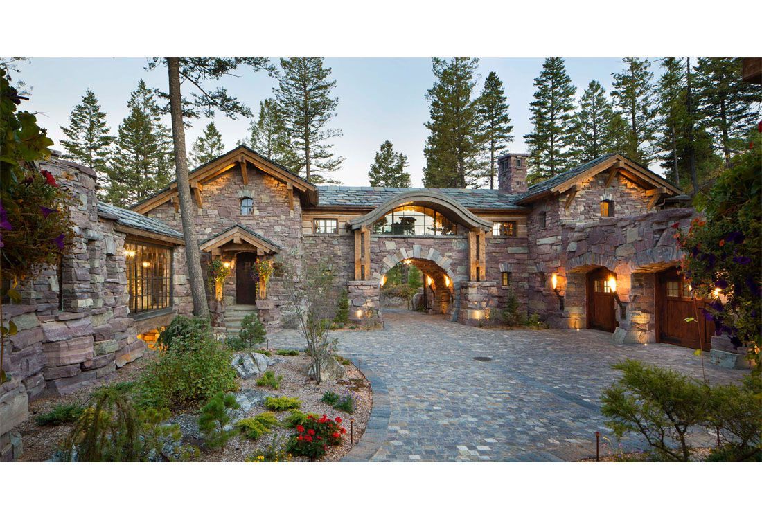 A large stone house with a driveway leading to it is surrounded by trees.
