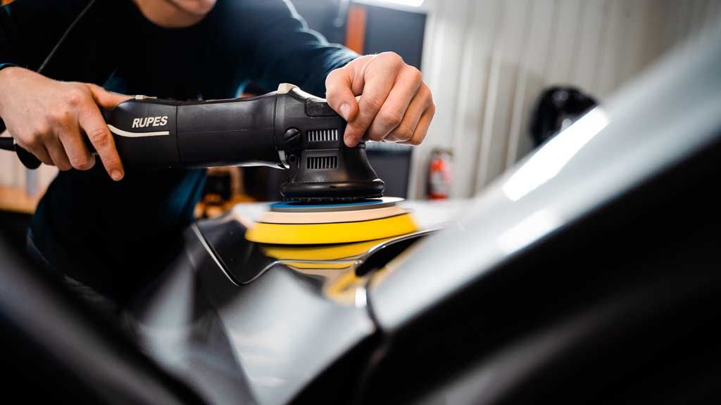 Person using a power polisher on a black car, polishing the vehicle's glossy surface.