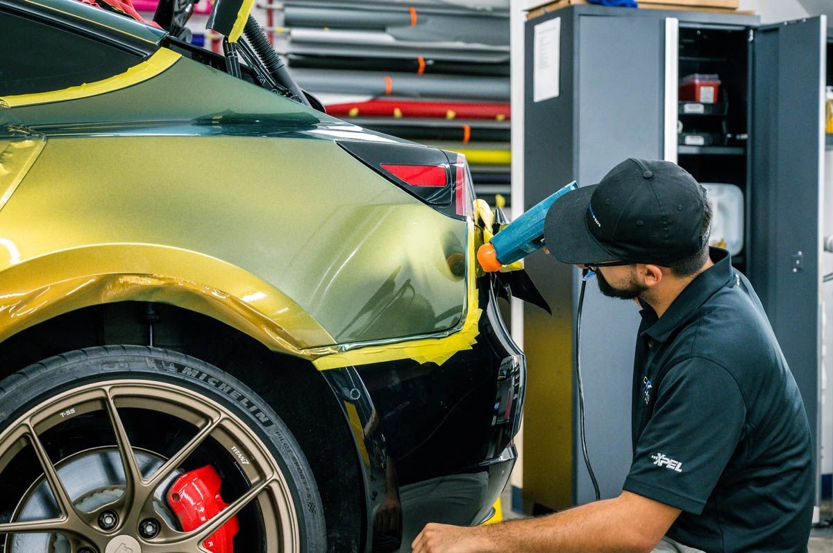 A person using a heat gun to apply a gold-colored wrap to a black car's rear bumper.