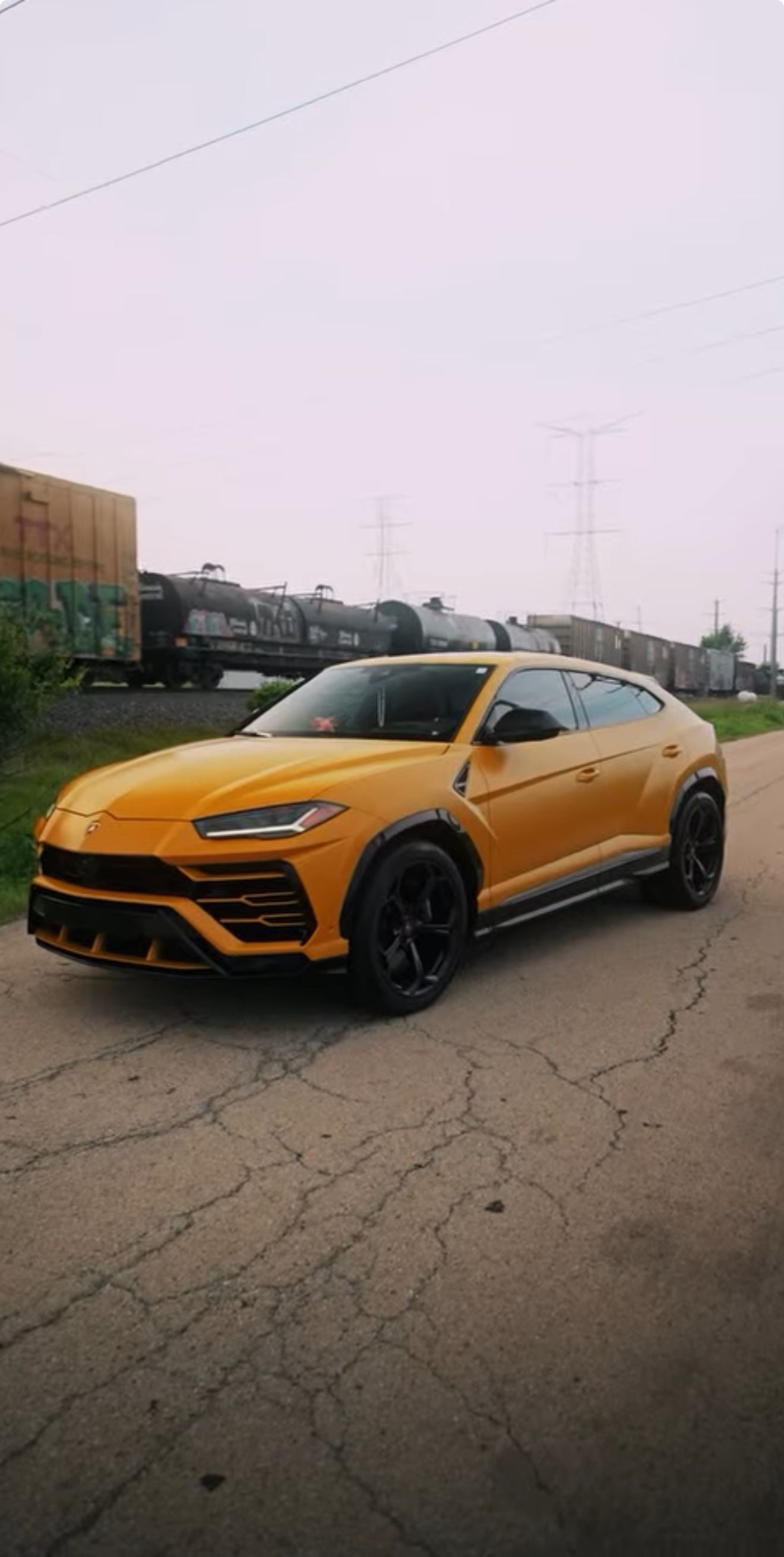 Yellow Lamborghini Urus parked on a cracked road, train in the background under an overcast sky.