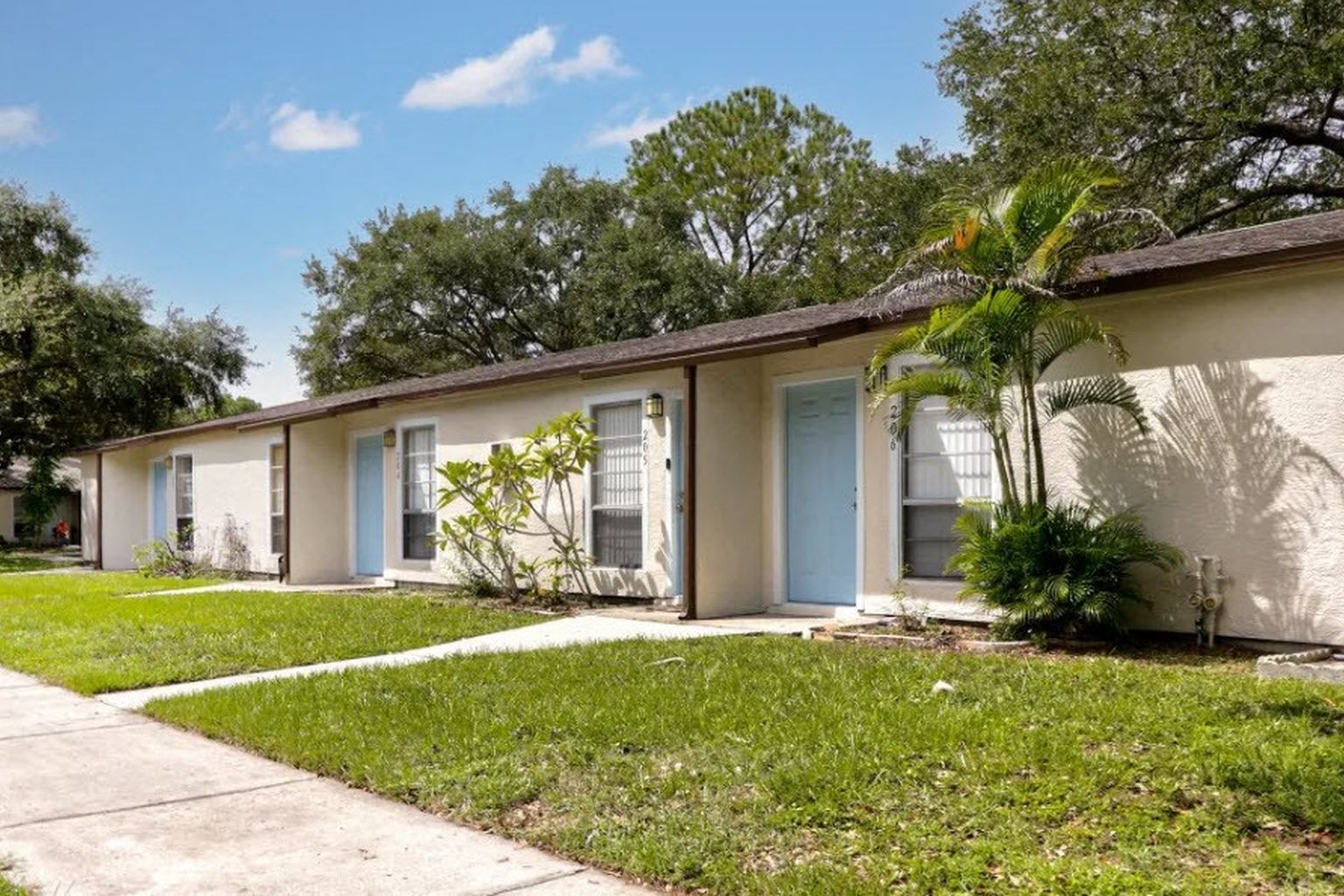 Row of beige apartments with light blue doors and windows, green lawn, and sidewalk on sunny day.