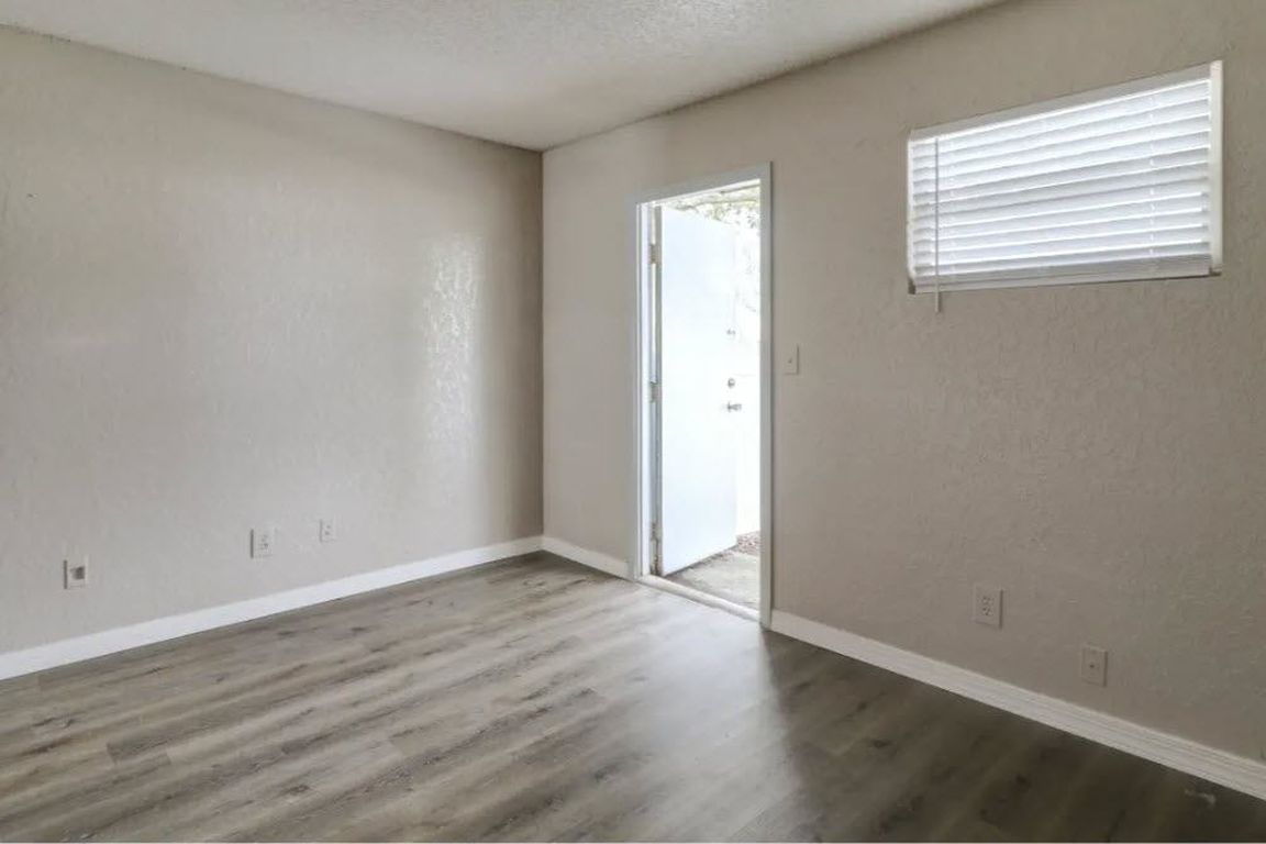 Empty room with grey walls, open white door, and window with blinds; gray wood-look floor.