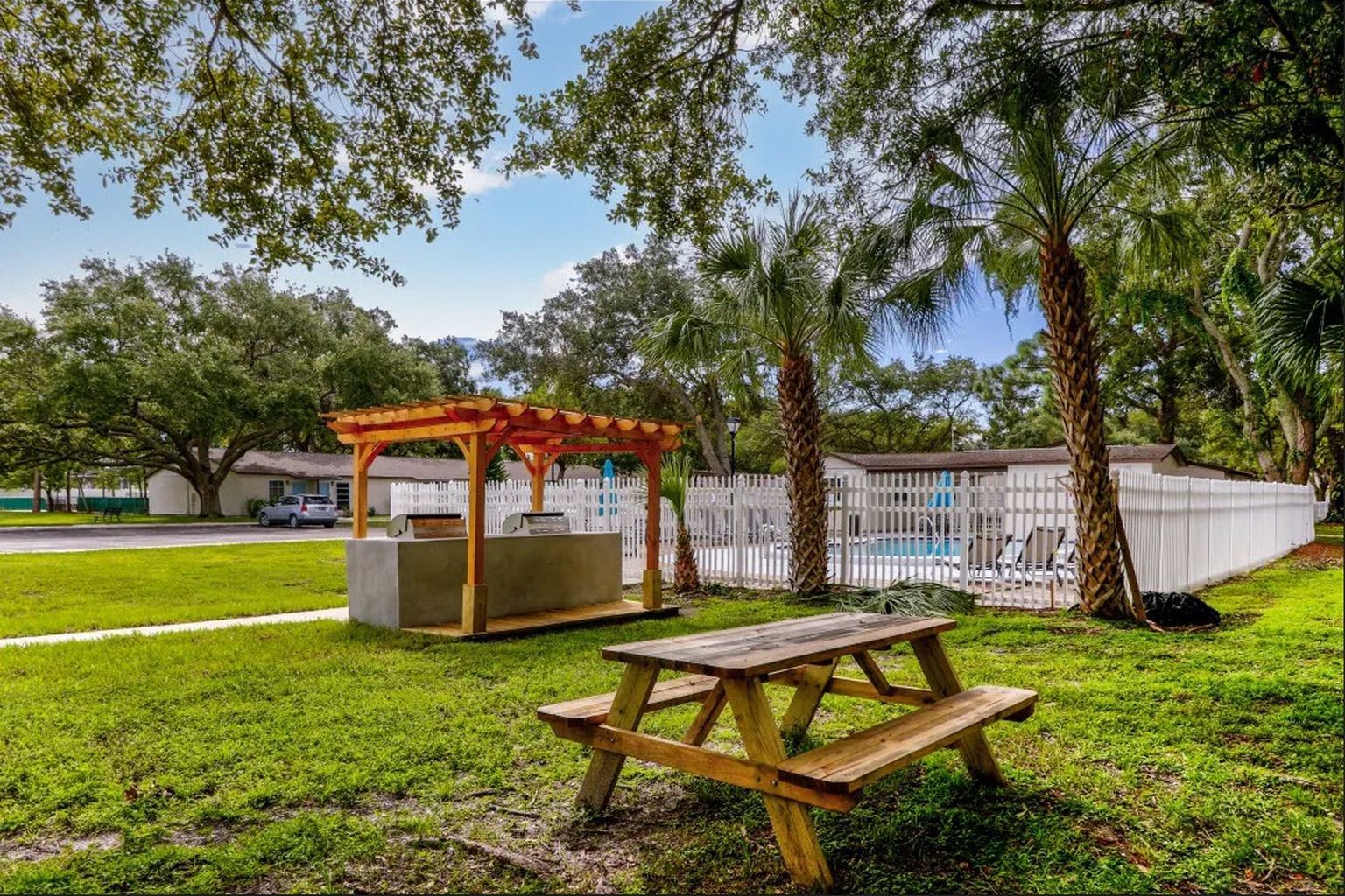 Picnic table in a grassy area, with a wooden pergola structure and a swimming pool in the background.