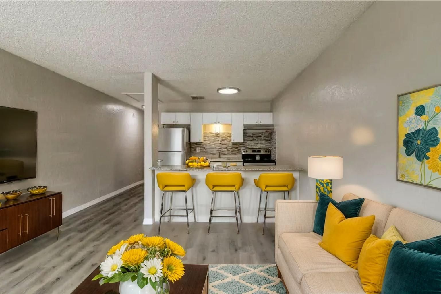 Living room with a kitchen in view, yellow accents, and a floral painting on the wall.
