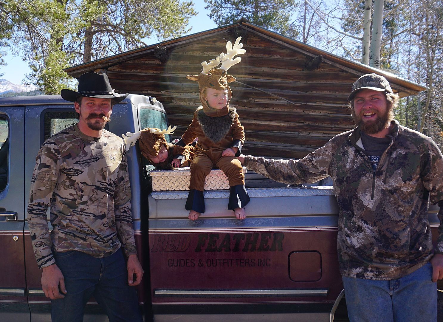 Two men are standing next to a truck with a child in a reindeer costume on the back.