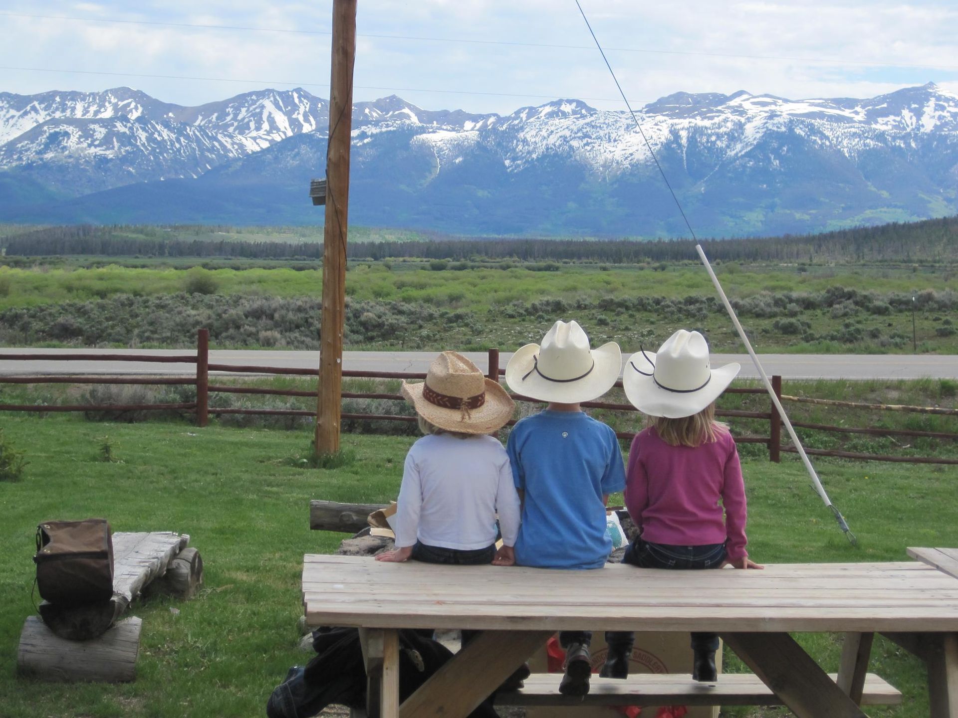 Three children wearing cowboy hats are sitting at a picnic table looking at the mountains