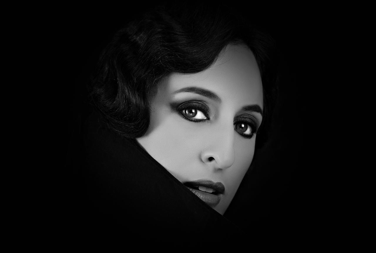 Woman with dark hair and dramatic eye makeup, emerging from a dark background.