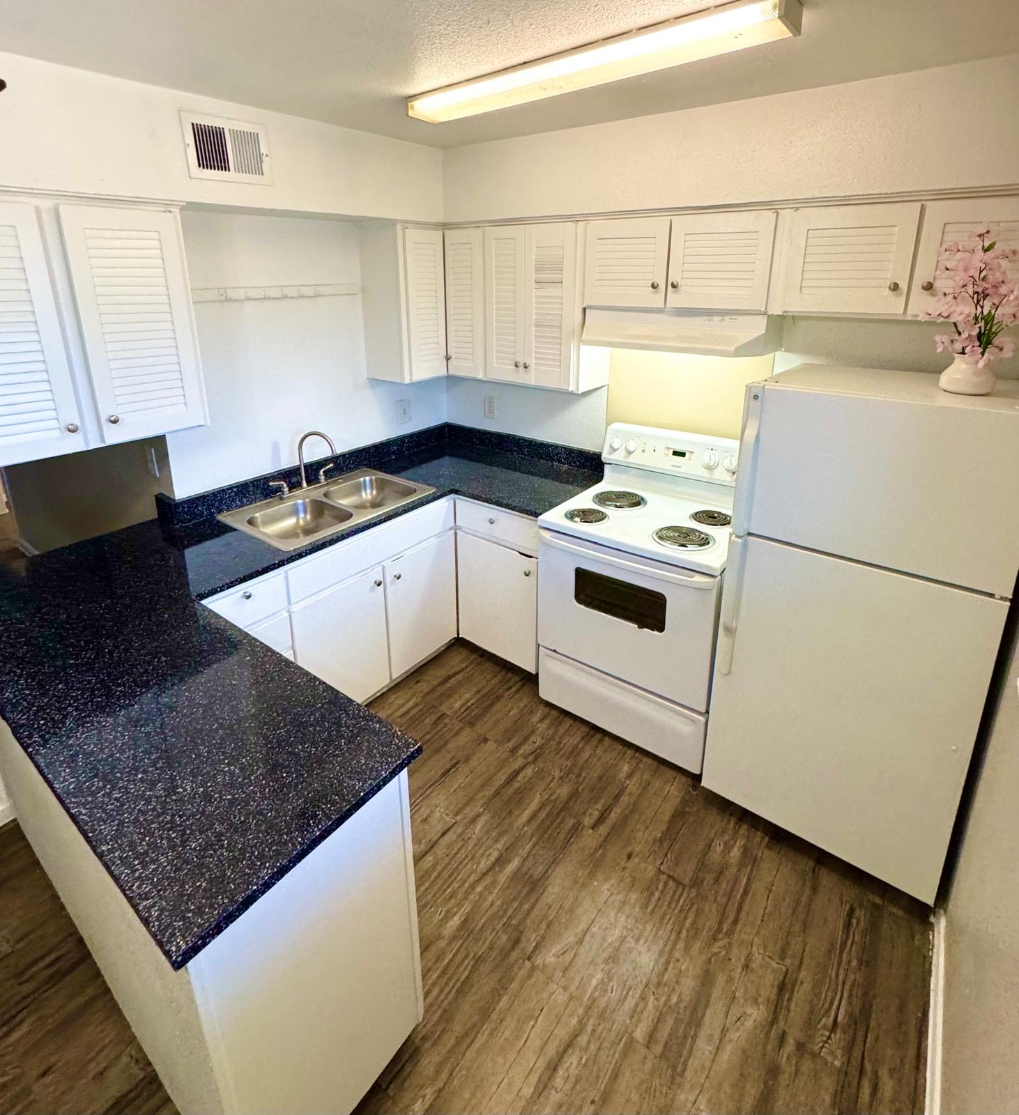Small, bright kitchen with white cabinets, black countertops, and white appliances. Wood-look flooring.