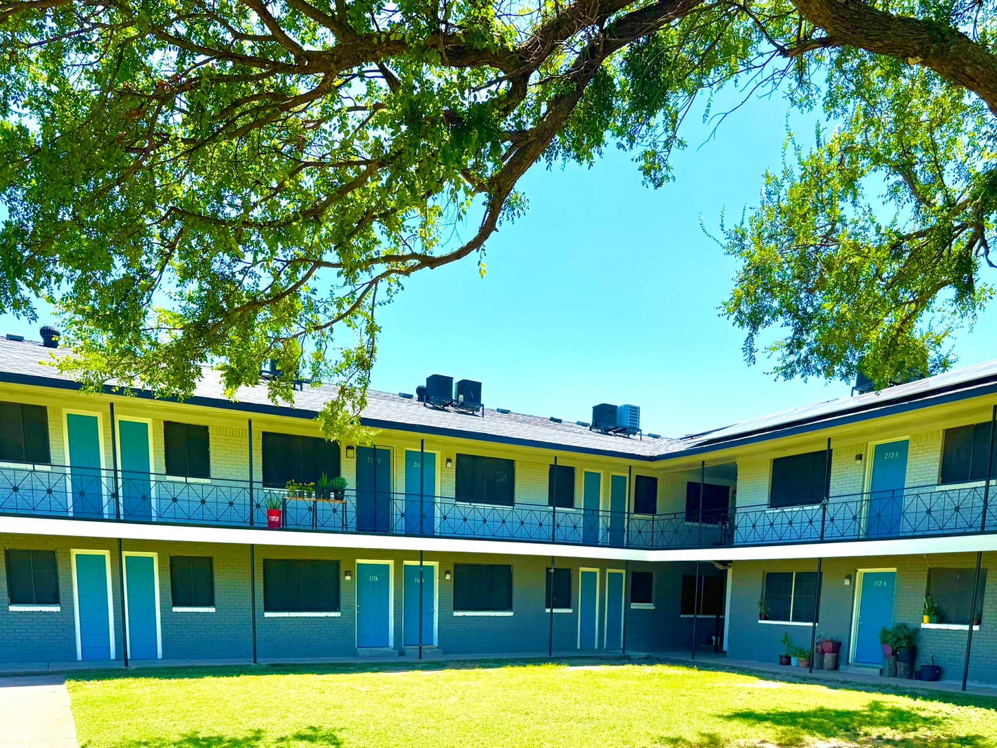 Apartment complex with blue doors, gray walls, and a central grassy courtyard shaded by a large tree under a clear blue sky.