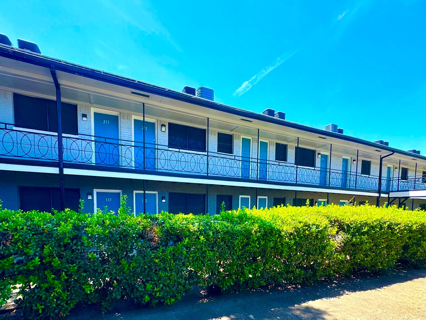 A two-story motel with light blue doors and decorative black ironwork, set against a clear blue sky and a row of green bushes.