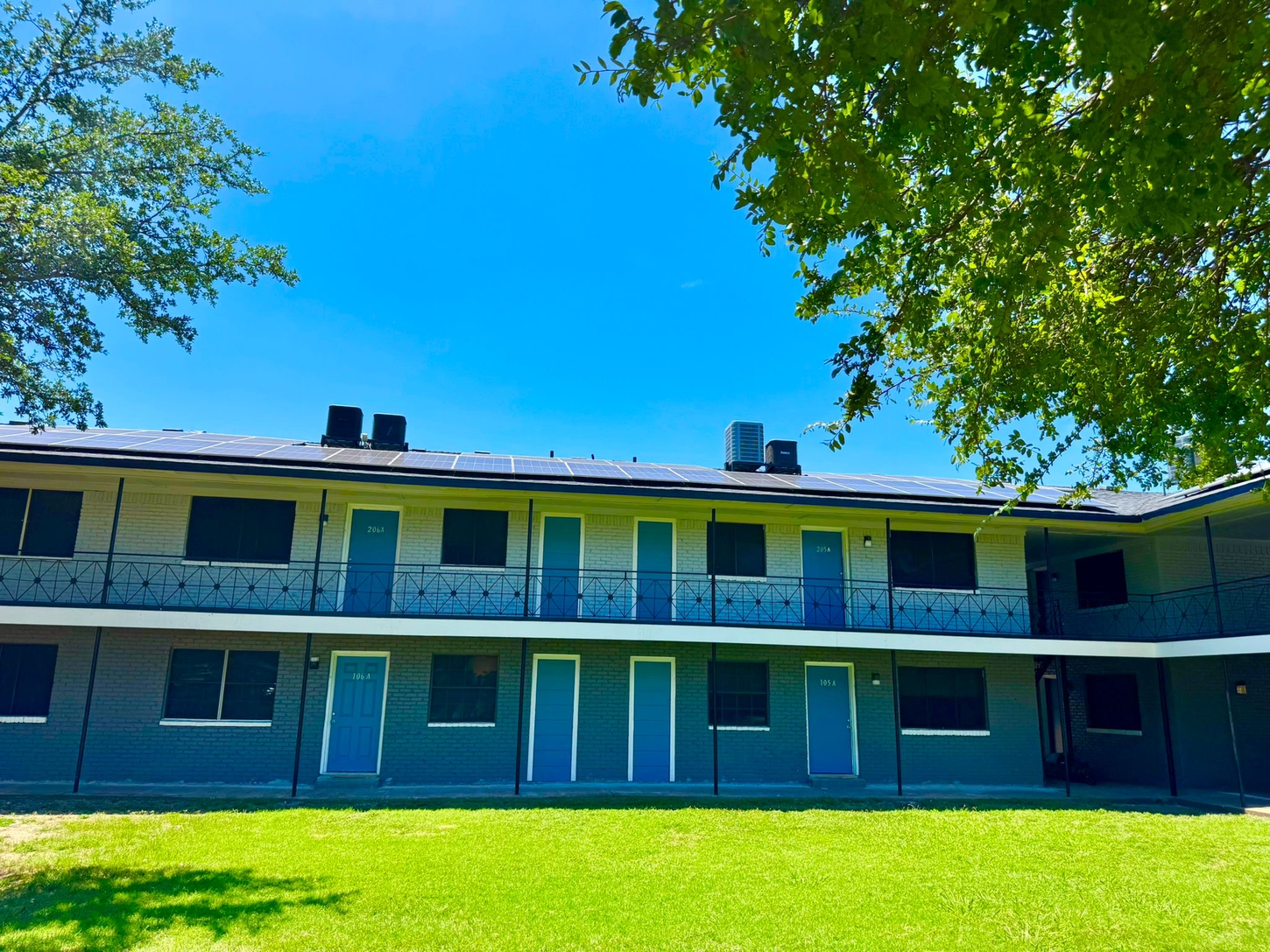 Two-story apartment building with blue doors and gray walls under a bright blue sky. Green grass in the foreground.