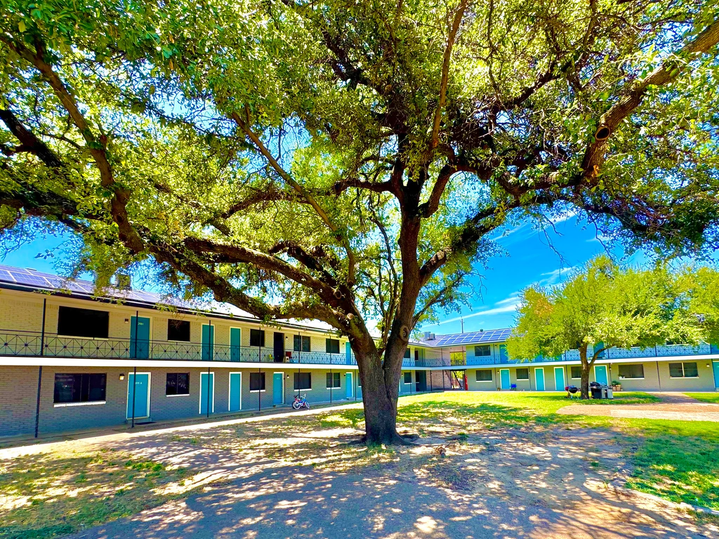 A two-story motel with turquoise doors and a large tree in the center courtyard under a bright blue sky.