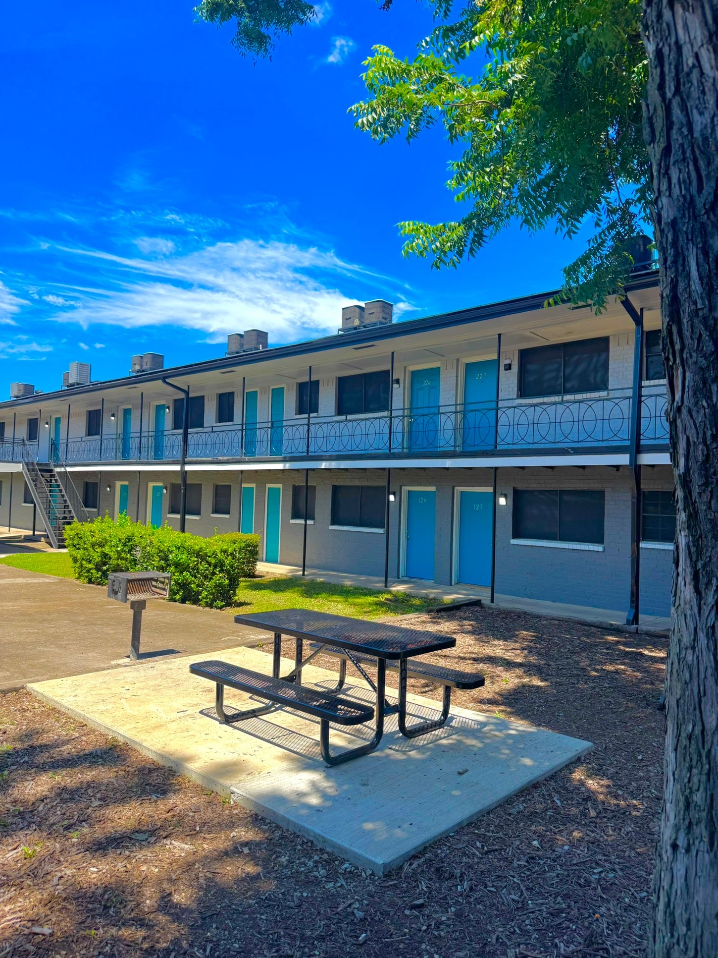 Two-story apartment building with blue doors, picnic table in front. Sunny day, blue sky, tree to the right.