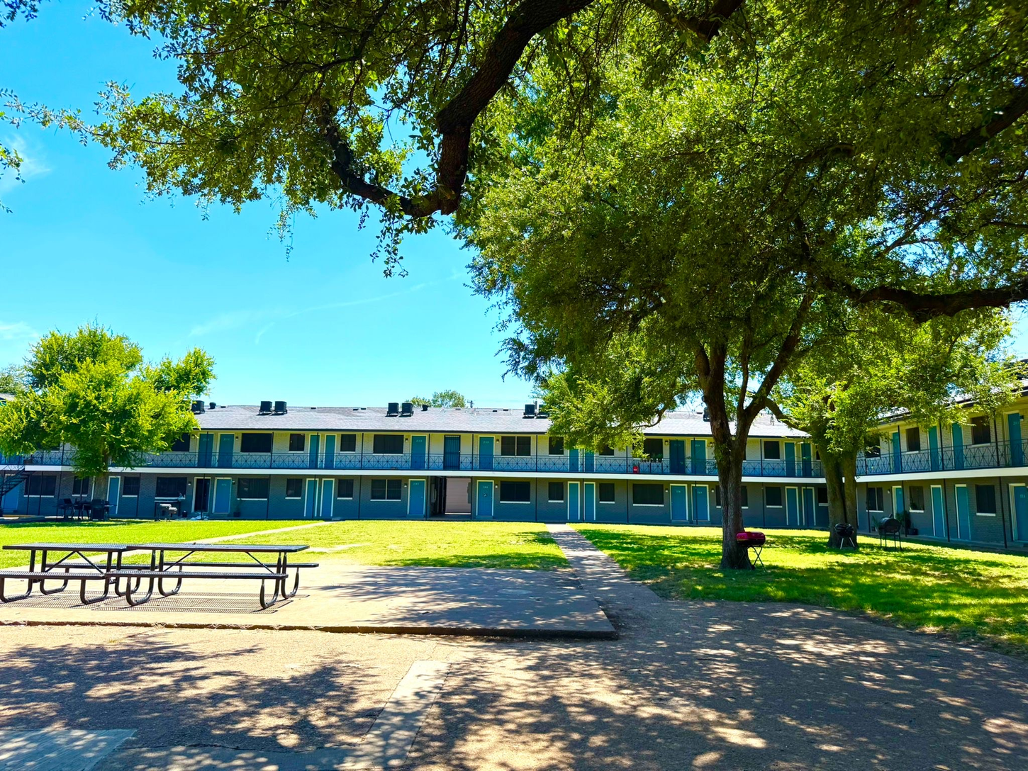 Exterior of a two-story building with teal doors and windows. A picnic table sits on a concrete area in front of it, shaded by trees.