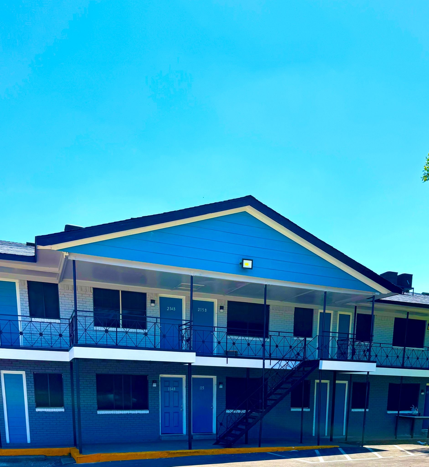 Two-story motel with blue trim and doors under a bright blue sky. A black staircase leads to the second floor.