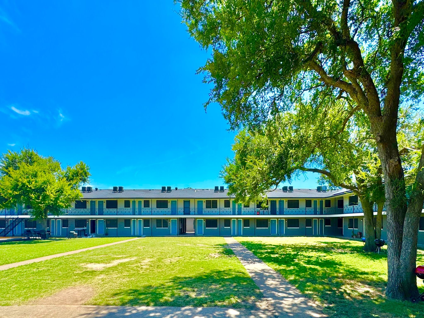 Two-story apartment building with blue doors and a grassy courtyard under a bright blue sky.
