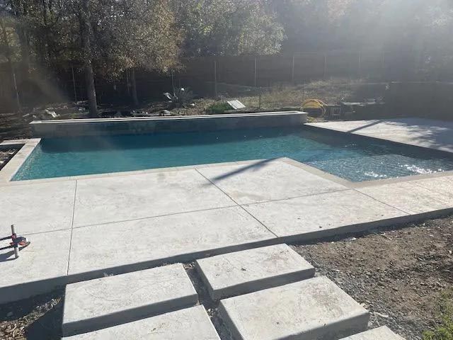 A large swimming pool is surrounded by concrete steps in a backyard.