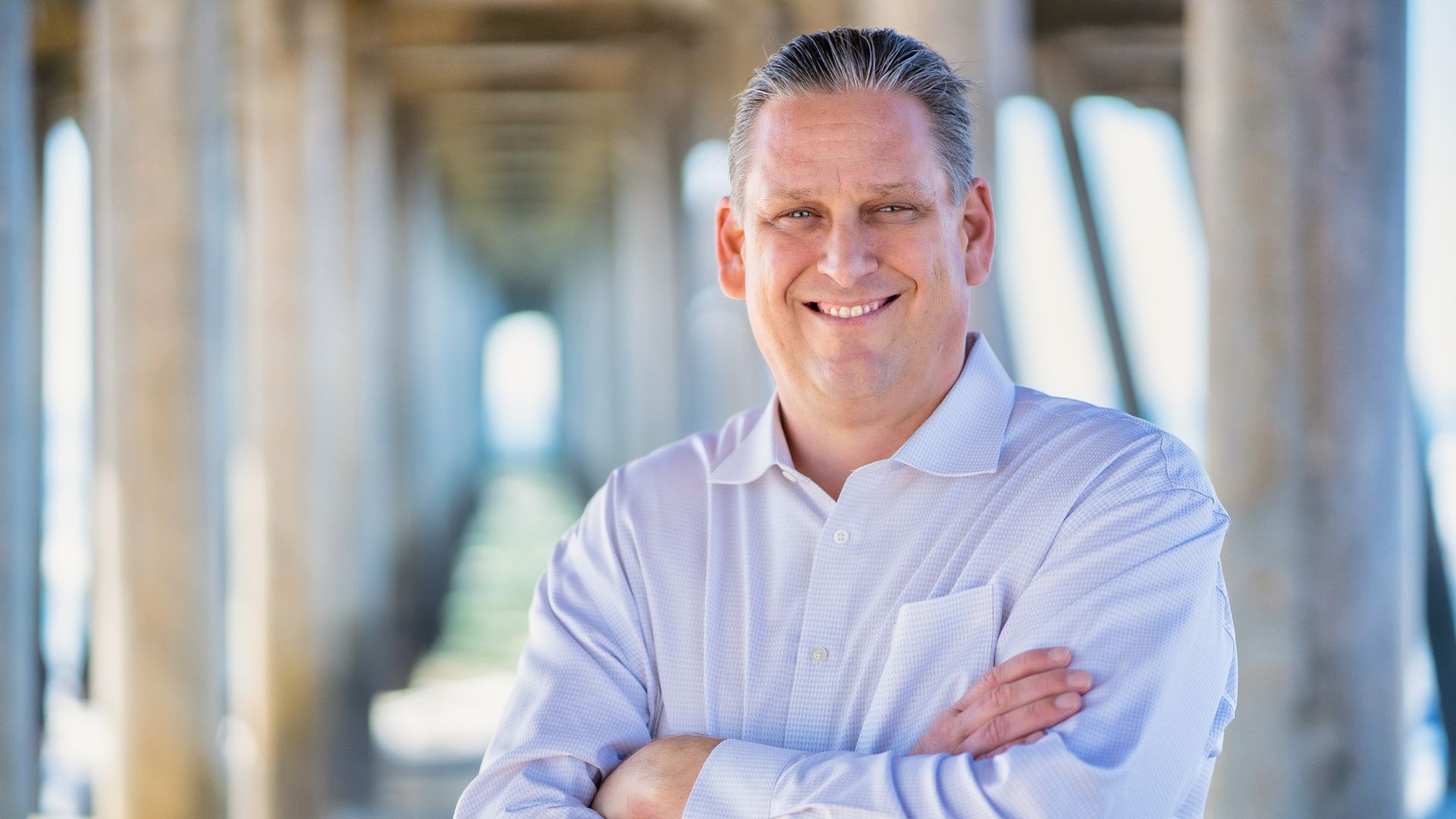 A man in a white shirt is standing under a pier with his arms crossed and smiling. Tony Strickland