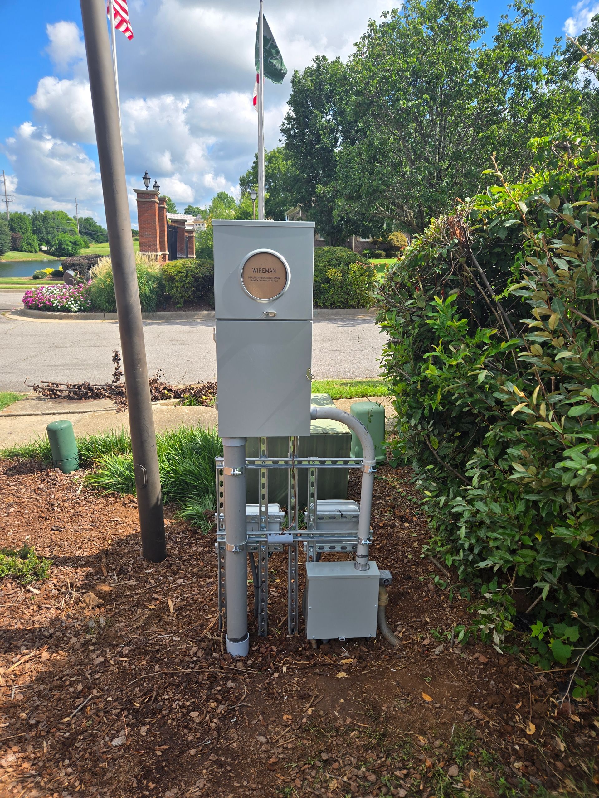 Gray electrical box and conduit near flags, with bushes and a cemetery in the background.