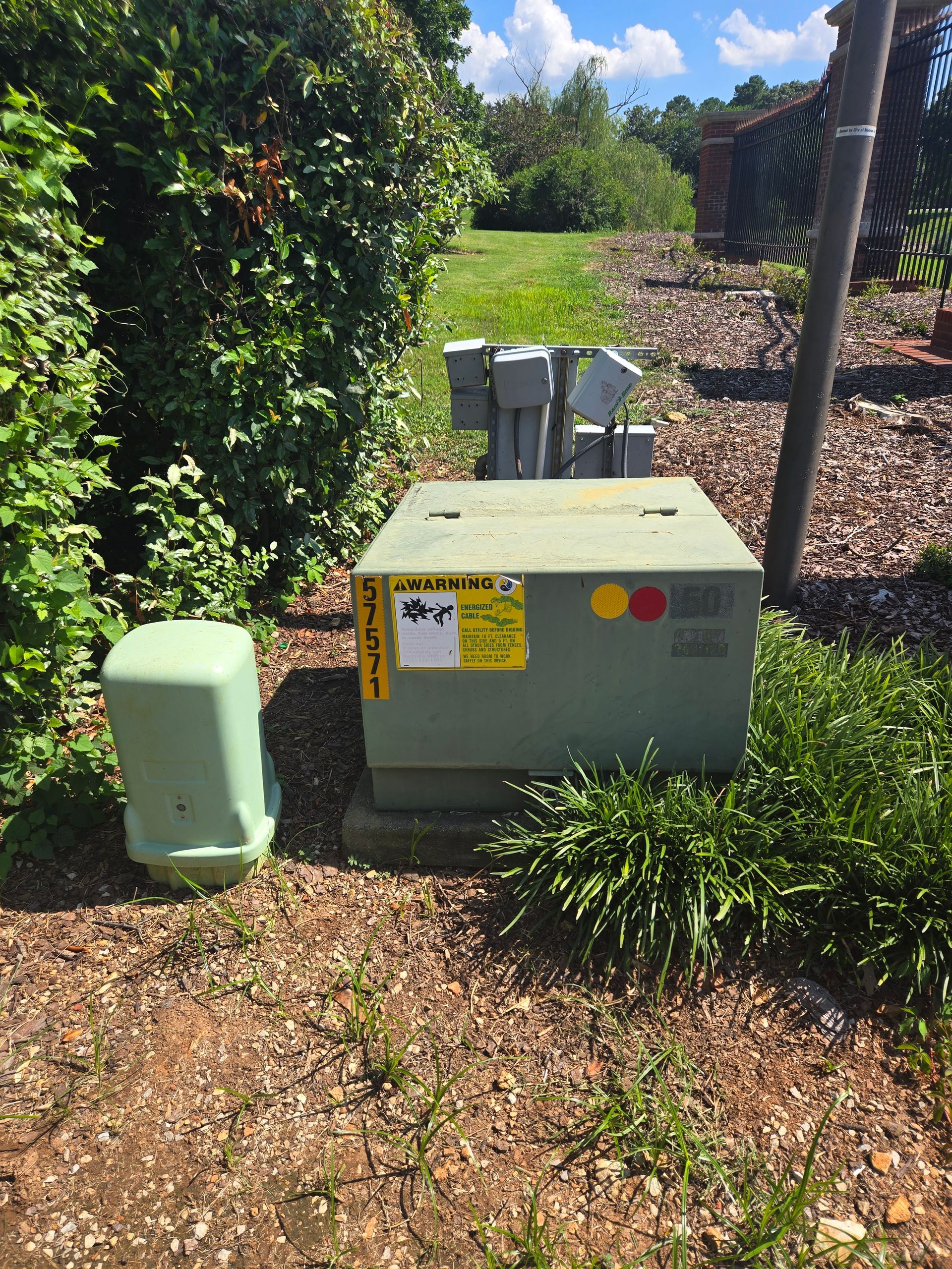 Green utility box with warning labels next to a green electrical enclosure and a fence in an outdoor setting.