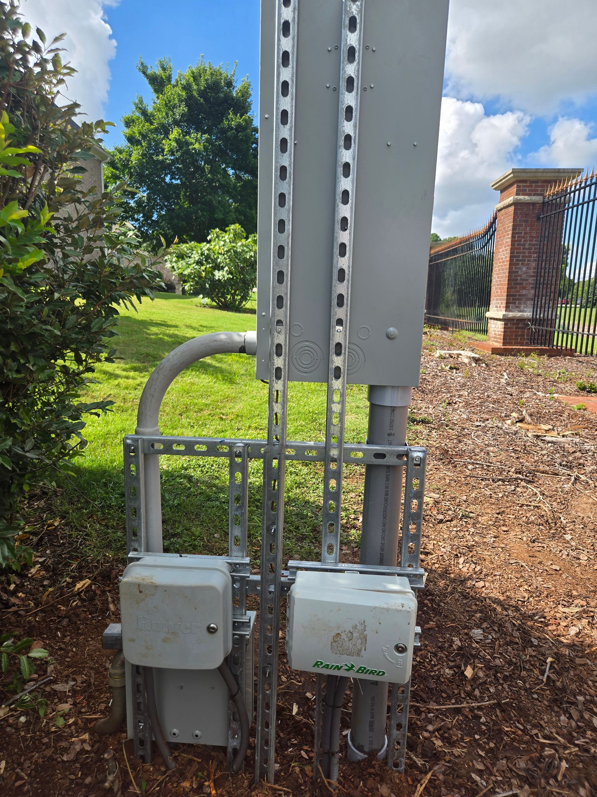 Outdoor electrical boxes mounted on a pole, with metal supports and conduits, in a grassy area near a brick fence.