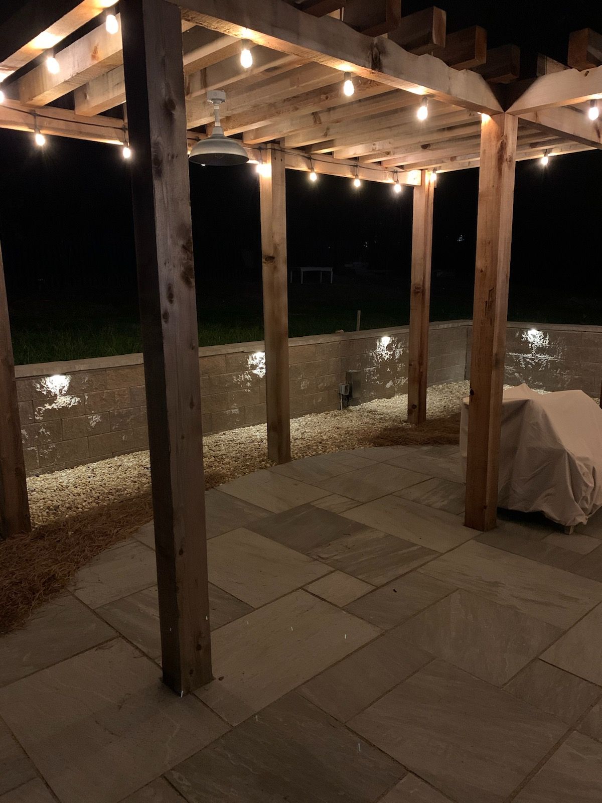 Patio with wooden pergola lit by string lights at night. Stone walls and flooring.