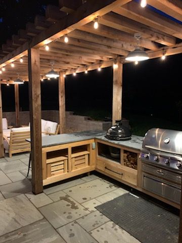 Outdoor kitchen at night under a pergola with string lights, built-in grill, and wooden cabinets.