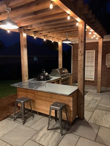 Outdoor kitchen with a wooden pergola and string lights. Has a grill, countertop, and two metal stools.