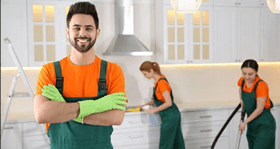 Man in green and orange uniform smiles at the camera in a kitchen while two others clean.