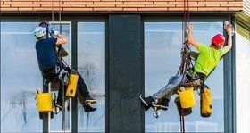 Two window washers suspended on ropes cleaning a tall building's windows; one in blue shirt, one in green.