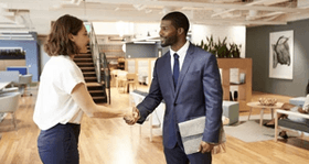 Woman and man shaking hands in a modern office. Man holds a laptop; both smile.