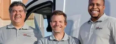 Three people smiling, wearing gray polo shirts. They are standing in front of a vehicle.
