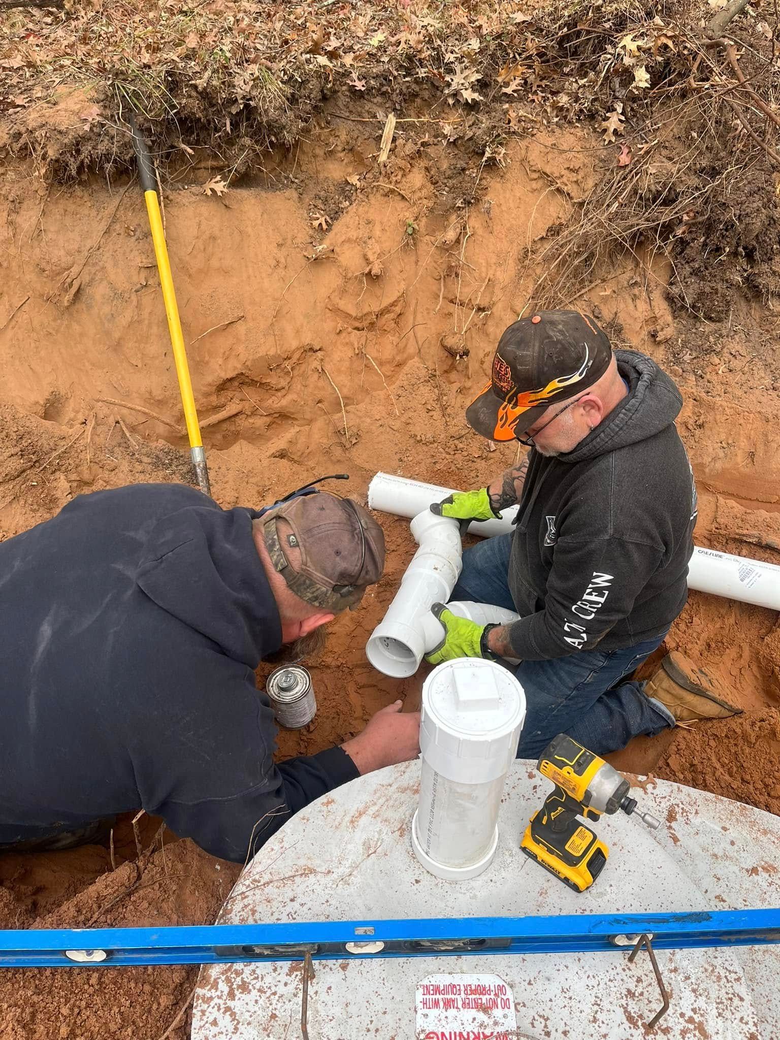 Two workers in a dirt trench install PVC pipes onto a concrete septic tank riser, using a drill and a level.