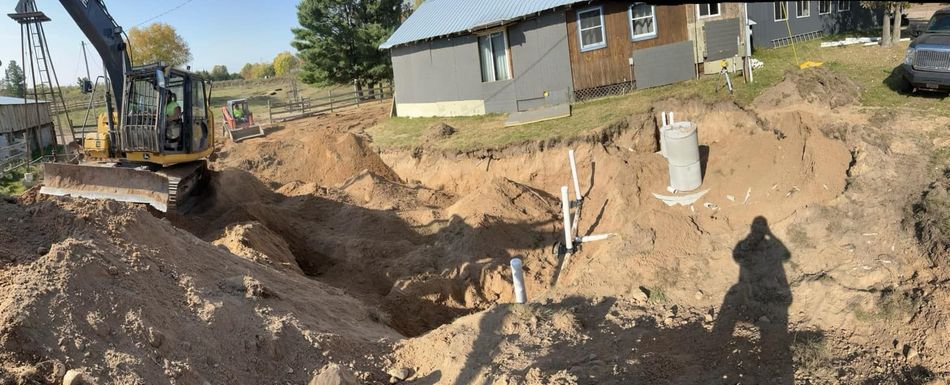An excavator digs a large trench next to a house with exposed pipes and a concrete septic riser visible in the dirt.