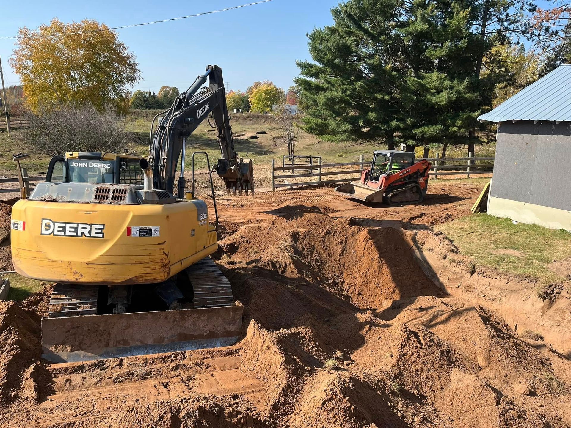 A yellow John Deere excavator and an orange skid steer working on an excavated dirt site near a building.