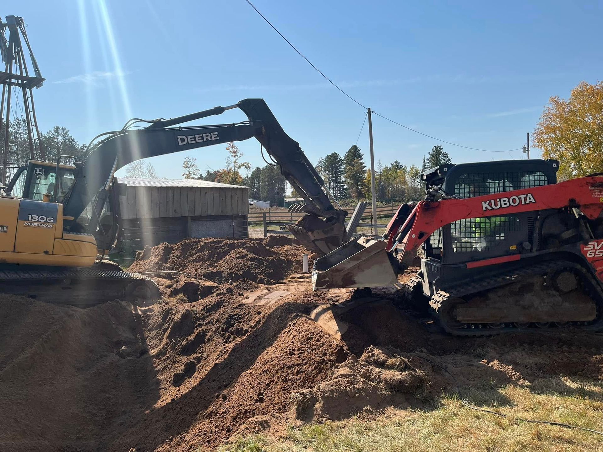 A yellow excavator and a red Kubota track loader working together on a mound of dirt at an outdoor construction site.