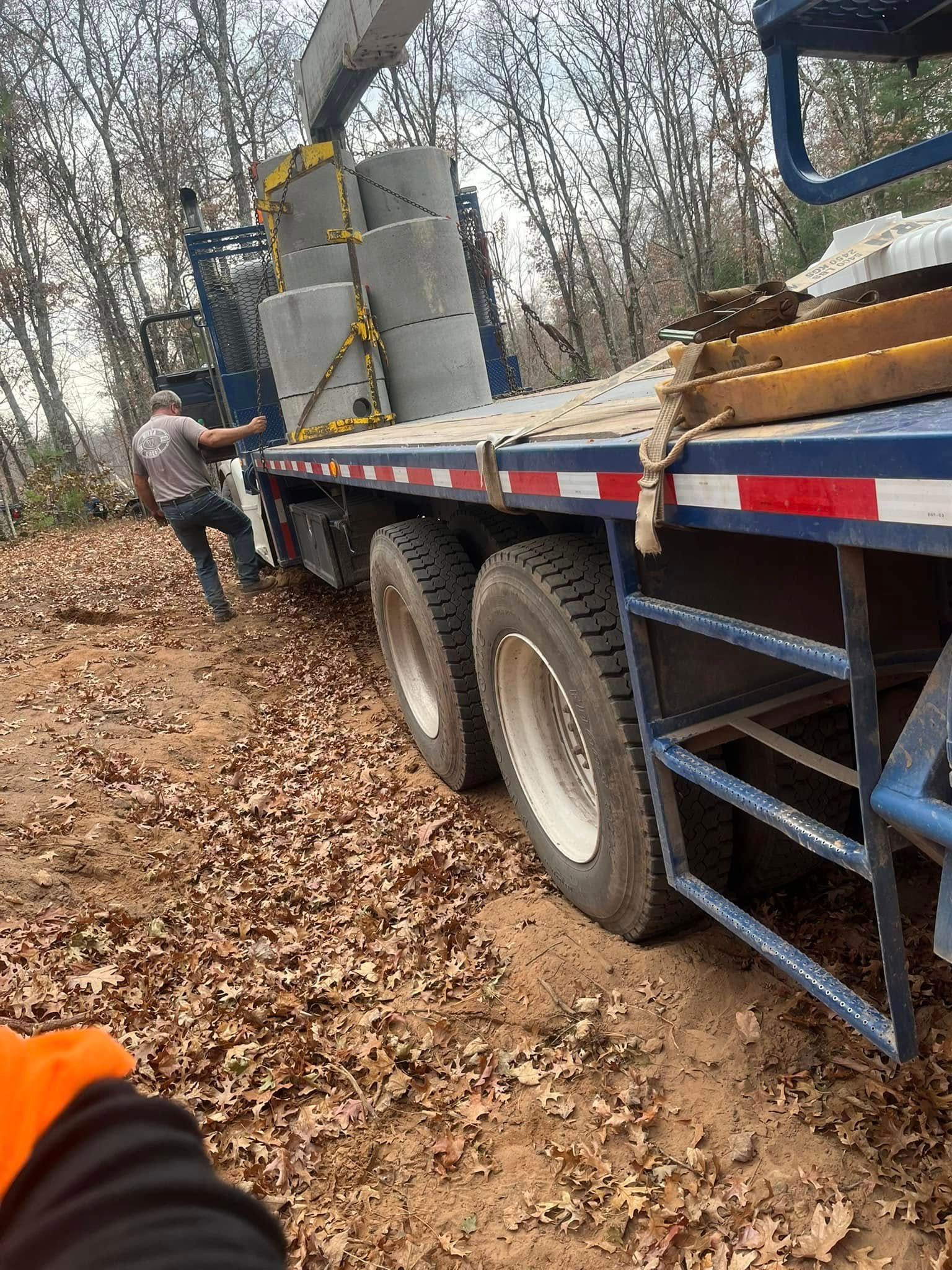 A worker stands next to a flatbed truck loaded with large concrete pipes parked on a forest floor covered in autumn leaves.