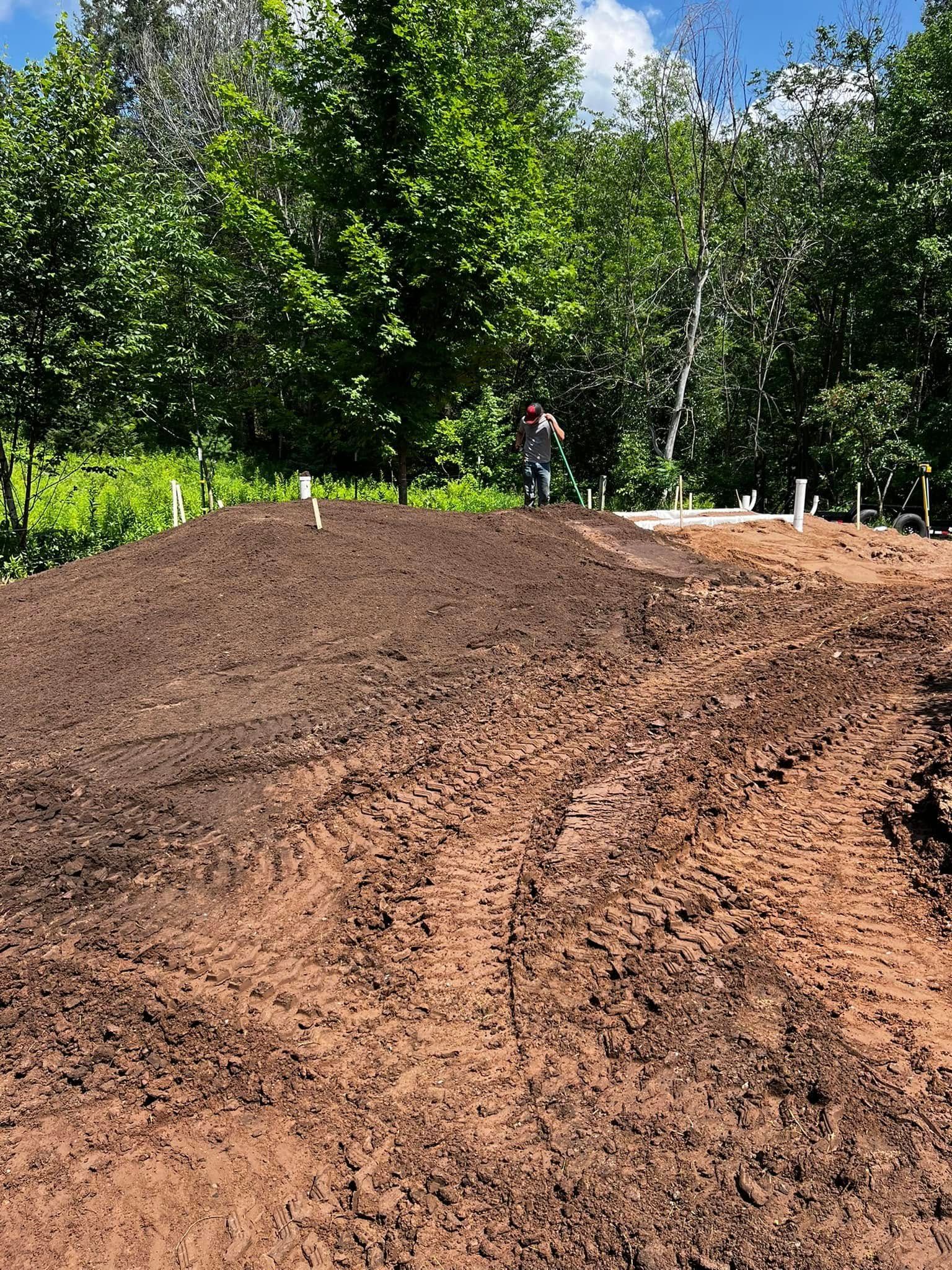A person walks along a large, freshly excavated dirt mound in front of a line of trees on a sunny day.