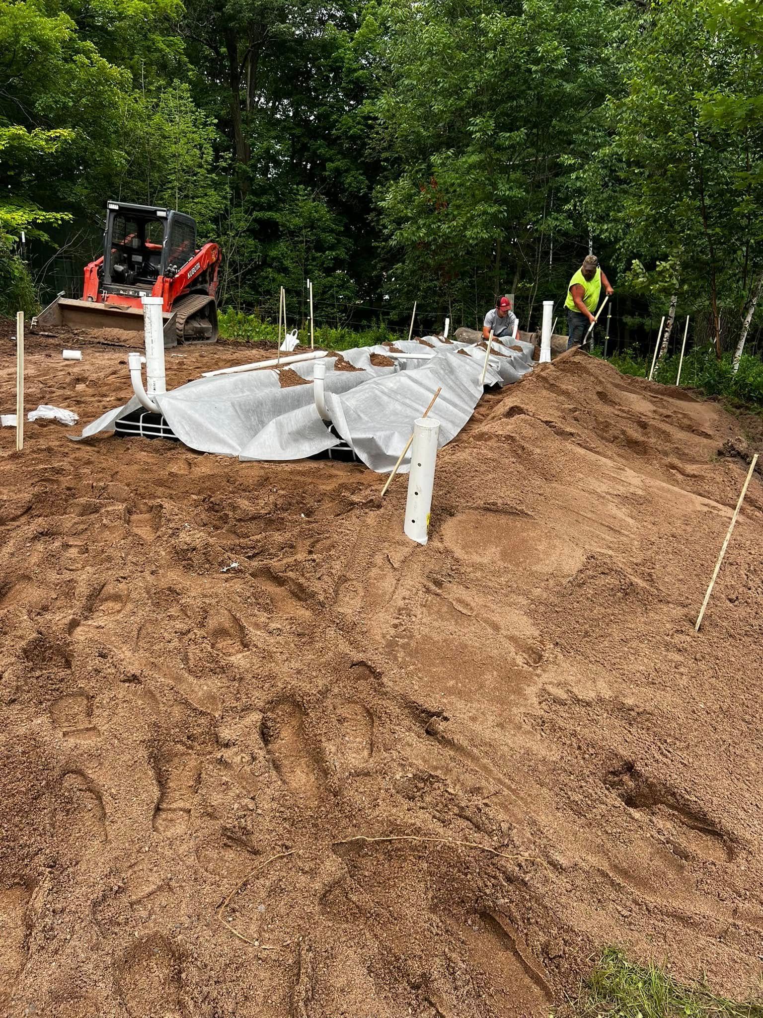 Workers install a residential septic system drain field, featuring white pipes and grey fabric cover in a wooded area.