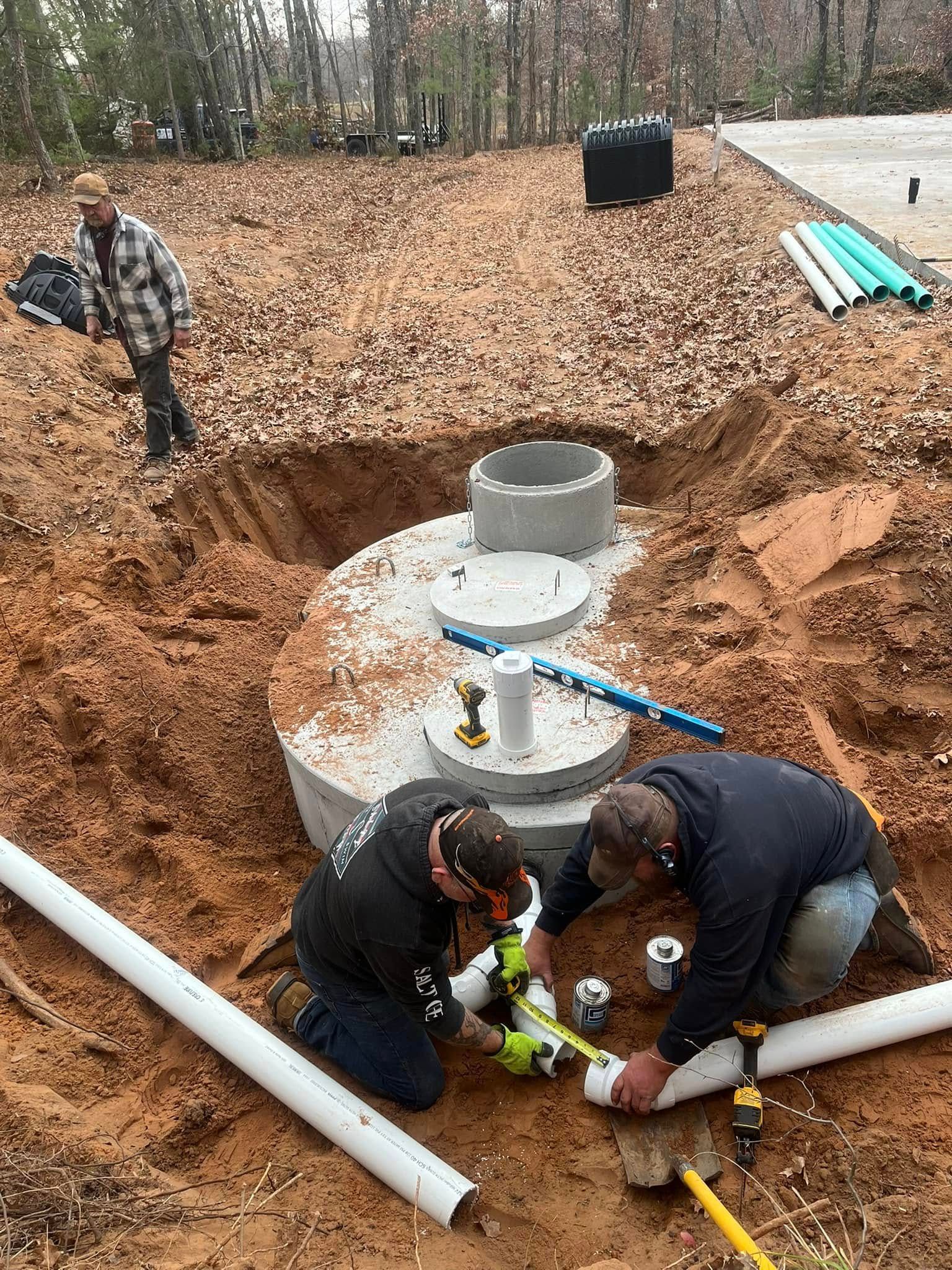 Two workers in a construction site install PVC piping into a concrete septic tank set in a dirt excavation.