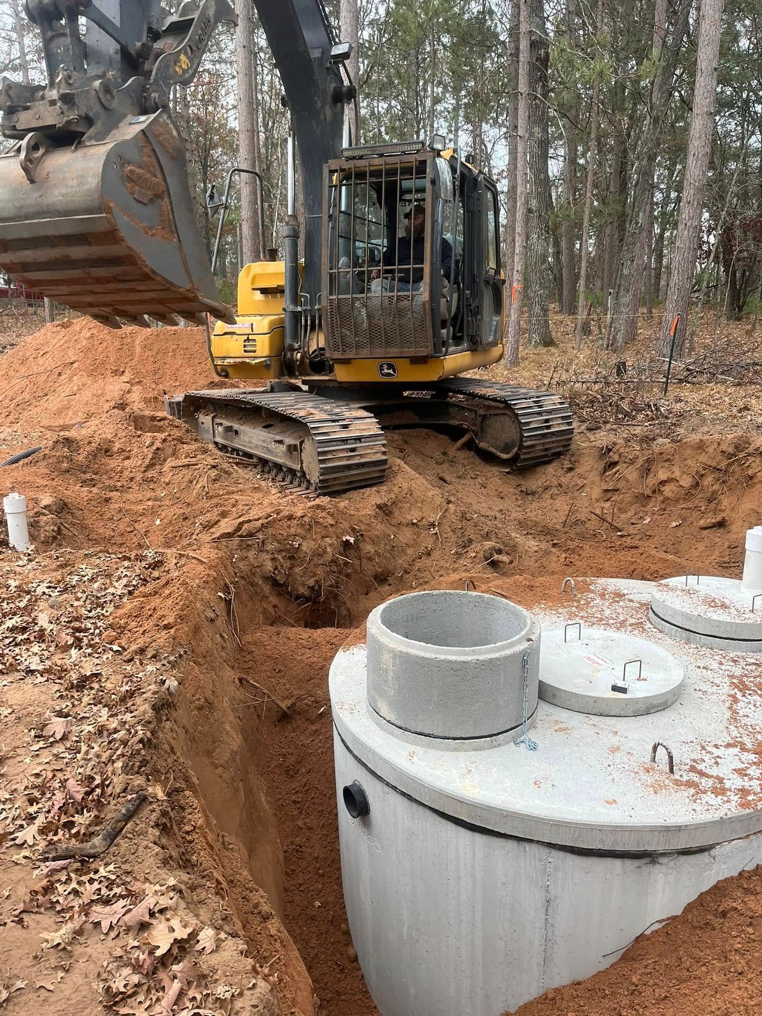 A yellow excavator sits in a wooded area next to a freshly installed concrete septic tank in an excavated trench.