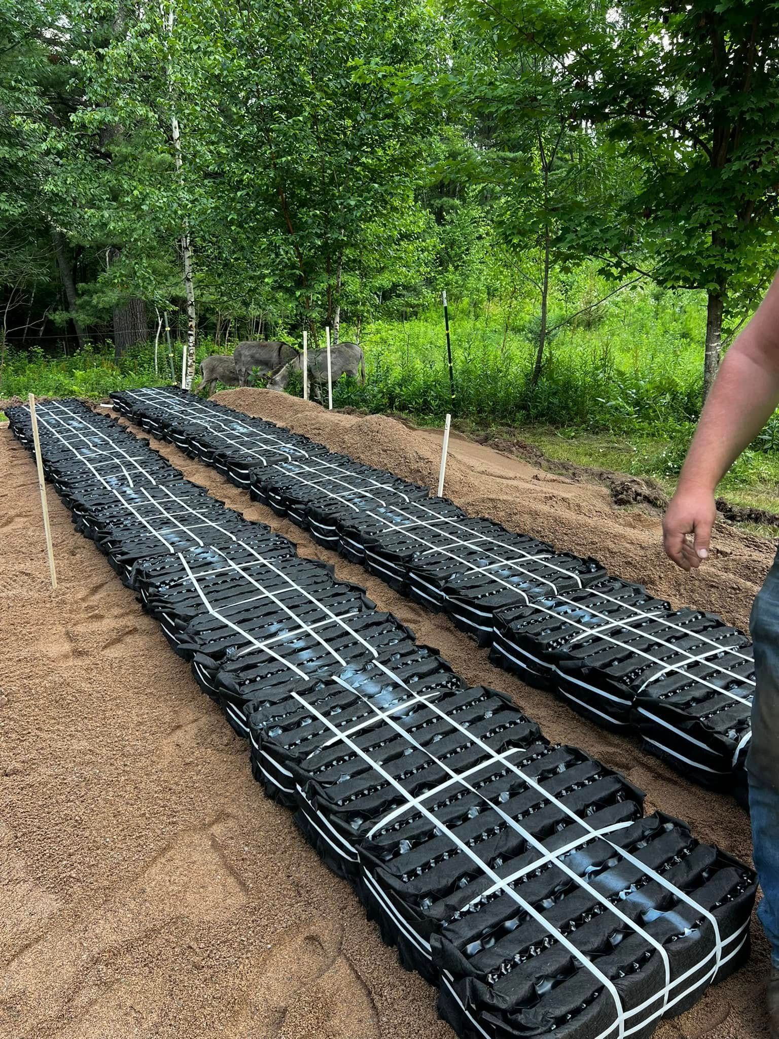 Two rows of stacked, black plastic drainage pipe segments secured with white straps on a prepared dirt bed in a forest.