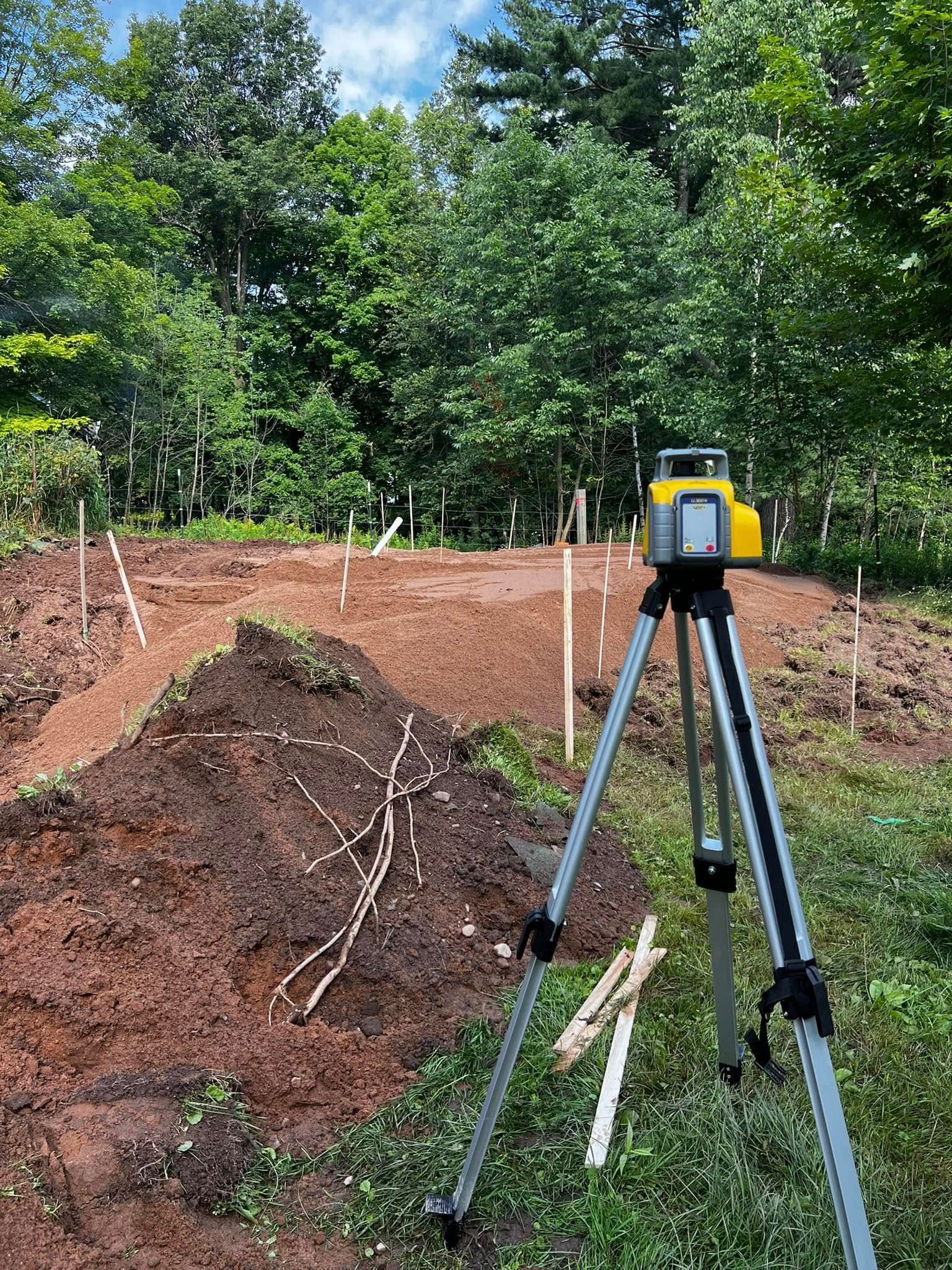 A yellow laser level on a tripod stands at a construction site with mounds of reddish dirt and wooden survey stakes.