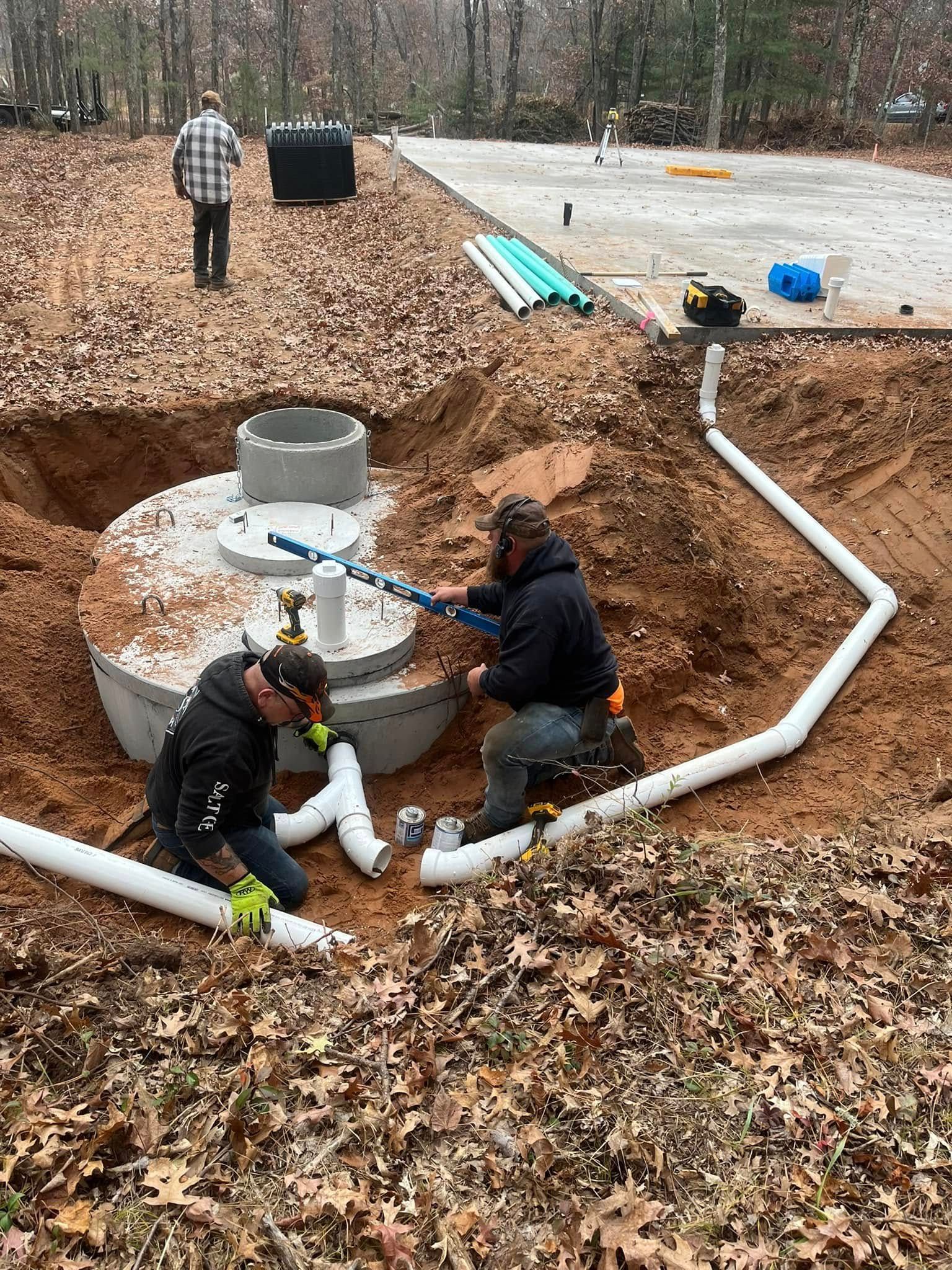 Two workers install white PVC piping to a concrete septic tank in an outdoor excavation site.