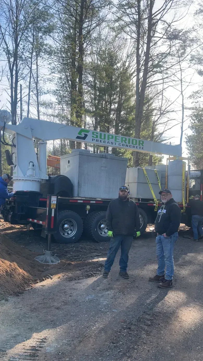 Two workers stand in front of a flatbed truck carrying a large concrete tank and a crane, parked on a wooded gravel road.